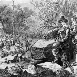 An 18th century engraving showing Puritans standing among some rocks looking down on settlers and Indigenous Americans dancing around a may-pole. The Puritans are all wearing armour as they are ready to clamp down on the spectacle below.  s 