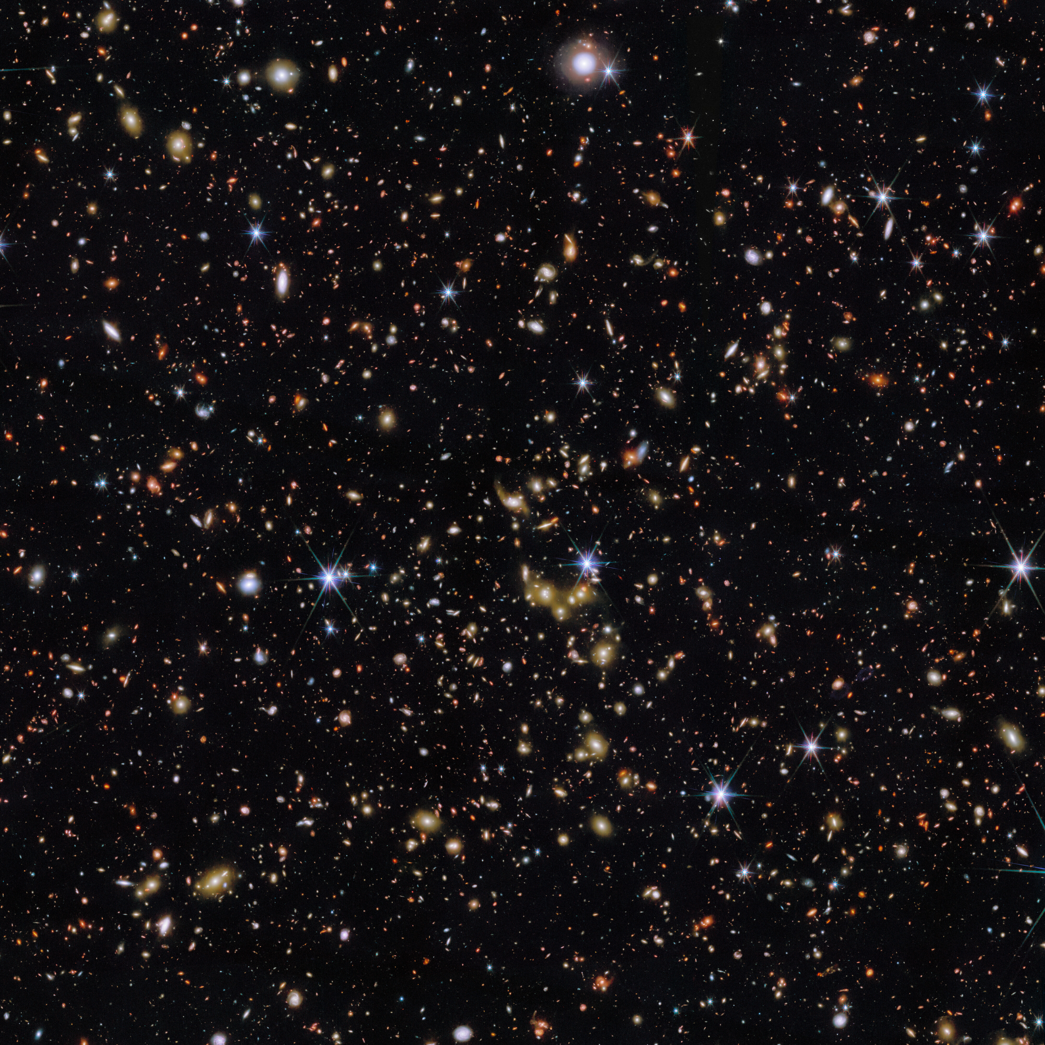 An area of deep space with thousands of galaxies in various shapes and sizes on a black background. Most are circles or ovals, with a few spirals. More distant galaxies are redder in colour and smaller, down to being mere dots, while closer galaxies are a bit larger and white or blueish. A few gold-coloured galaxies are bunched closely together in the centre. Bright stars surrounded by spikes lie in our galaxy. An area of deep space with thousands of galaxies in various shapes and sizes on a black background. Most are circles or ovals, with a few spirals. More distant galaxies are redder in colour and smaller, down to being mere dots, while closer galaxies are a bit larger and white or blueish. A few gold-coloured galaxies are bunched closely together in the centre. Bright stars surrounded by spikes lie in our galaxy.