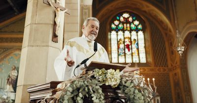 priest wearing white vestments preaching from the pulpit in a church; there is a stained glass window behind him and a crucifix, and the pulpit is decorated with flowers