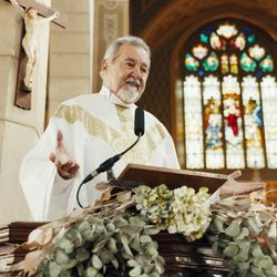 priest wearing white vestments preaching from the pulpit in a church; there is a stained glass window behind him and a crucifix, and the pulpit is decorated with flowers