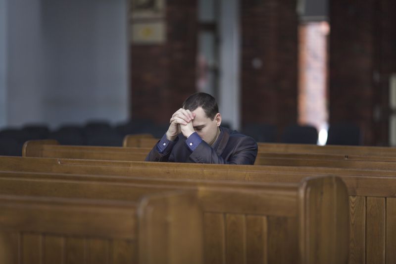 A man wearing a black jacket is resting his head against his hands as he prays in a church. The shot focuses on him sitting on his own among the pews. 