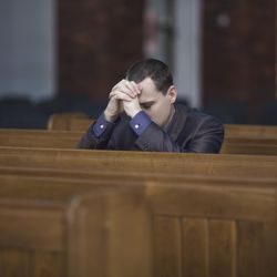 A man wearing a black jacket is resting his head against his hands as he prays in a church. The shot focuses on him sitting on his own among the pews. 