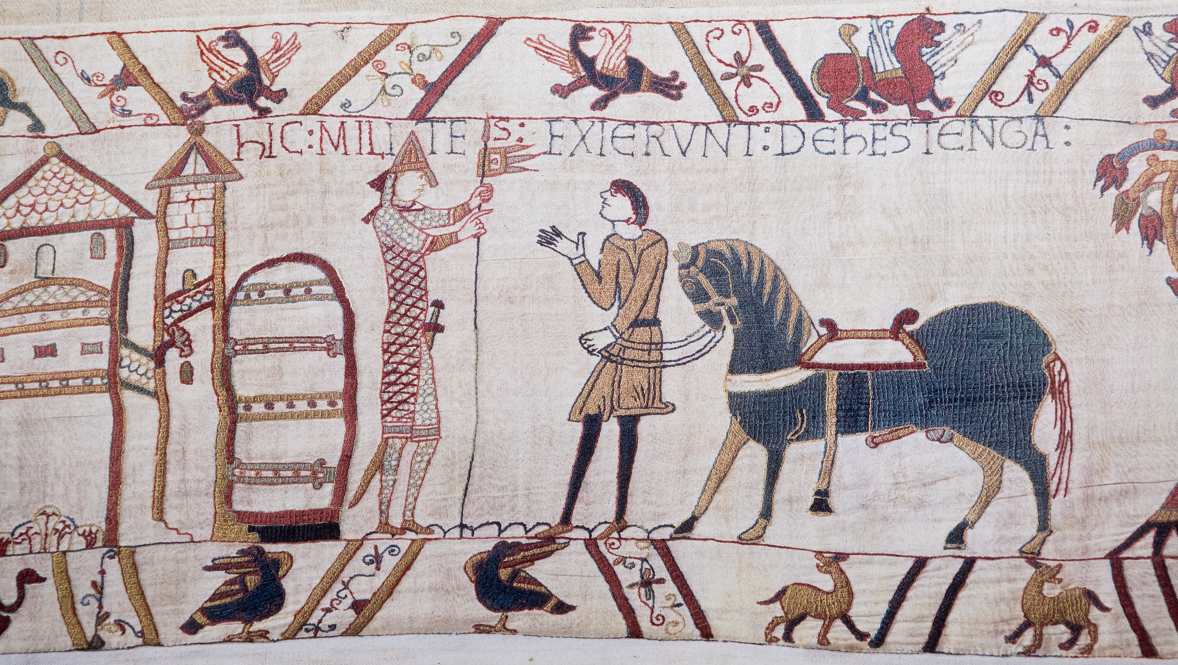 A scene showing Duke William leading his well endowed horse as he consults a male figure wearing armour, a helmet and holding a spear on the left of the image. The man has his back to a castle-like structure. The margins above and below the image show designs as well as strange bird-like creatures and what look like goats or similar animals. A scene showing Duke William leading his well endowed horse as he consults a male figure wearing armour, a helmet and holding a spear on the left of the image. The man has his back to a castle-like structure. The margins above and below the image show designs as well as strange bird-like creatures and what look like goats or similar animals.