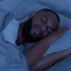 Portrait of happy young well-slept African American man lying in bed with closed eyes, resting in bedroom on the side in the dark night