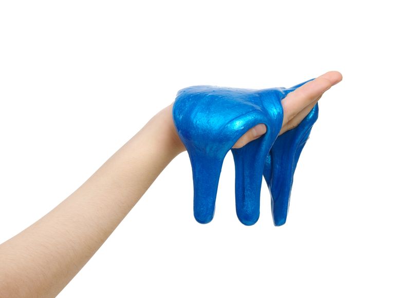 A close up of a person's hand holding blue slime in their hand. The slim is glooping over their palm and starting to drop to the the floor. The hand is set against a white background. 