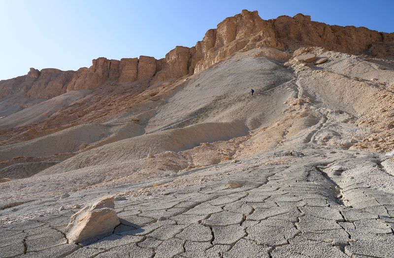 Male tourist in a hiking trail descending from a top of the Mount Zin or Har Zin in the Har Hahar nature reserve in Negev Desert. Unique landscape of the mount. Dried up cracked soil in the foreground