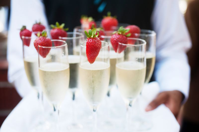 close up of tray being carried by a waiter, holding several flutes of champagne each with a strawberry wedged onto the top of the glass