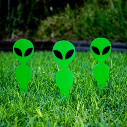 Five small typically alien-shaped lawn ornaments are standing in a V formation facing the camera. They are planted in the ground and probably about 4 to 5 inches tall. They are green and have large heads with black eyes. 