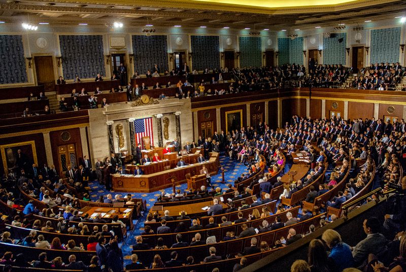 The photo shows the US Congress meeting and mingling on the house floor. The seating is arranged in the iconic semi-circular set up. The seats are filled with representatives, as is the upstairs balcony. 