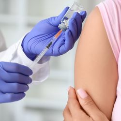 close up of doctor's hands as they draw up a vaccine in a syringe next to the patient's arm, with sleeve rolled up ready to get the shot