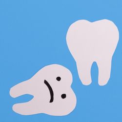 paper cutouts of three human teeth on a blue background, with an extra one lying on its side on the left with a sad face drawn on it