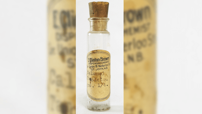 A photo of an old bottle of calomel ills. The bottle, which has a large cork stopper in its top is slim and clear, with a narrower but short neck. The bottle is empty and has a label on it saying "E Clinton Brown. Dispensing chemist. Union and Waterloo Street. St John, NB. Calomel tablets." The label is worn and browned with age.