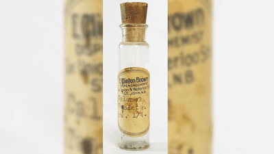 A photo of an old bottle of calomel ills. The bottle, which has a large cork stopper in its top is slim and clear, with a narrower but short neck. The bottle is empty and has a label on it saying "E Clinton Brown. Dispensing chemist. Union and Waterloo Street. St John, NB. Calomel tablets." The label is worn and browned with age.