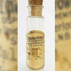 A photo of an old bottle of calomel ills. The bottle, which has a large cork stopper in its top is slim and clear, with a narrower but short neck. The bottle is empty and has a label on it saying "E Clinton Brown. Dispensing chemist. Union and Waterloo Street. St John, NB. Calomel tablets." The label is worn and browned with age.