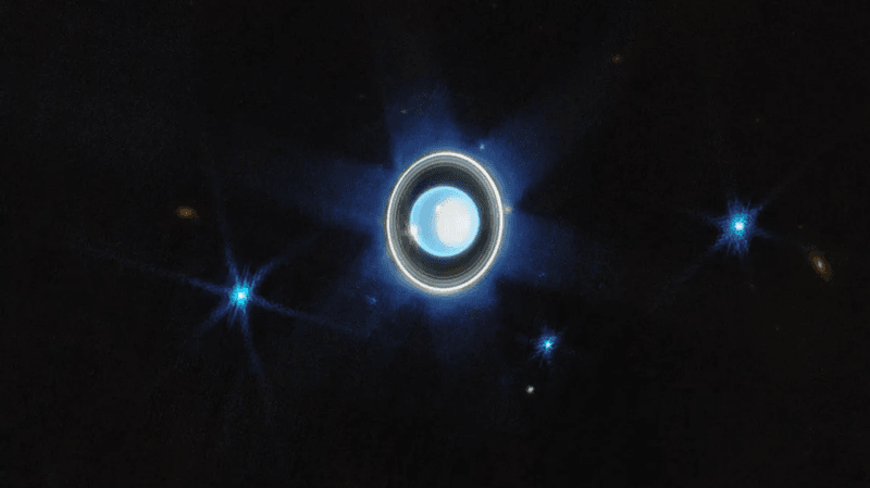 The planet Uranus is on a black background just left of centre. Just below the planet at the 7 o’clock position is a faint blue point labeled Puck. Brighter blue points at 8 o’clock, 5 o’clock, and 3 o’clock are labeled Ariel, Miranda, and Umbriel, respectively.